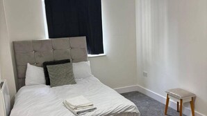 A neatly made bed with a grey headboard, a small bedside table, and a window with black curtains. - London Brigde Luxurious Spacious Apartment (London)