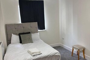 A neatly made bed with a grey headboard, a small bedside table, and a window with black curtains. - London Brigde Luxurious Spacious Apartment (London)