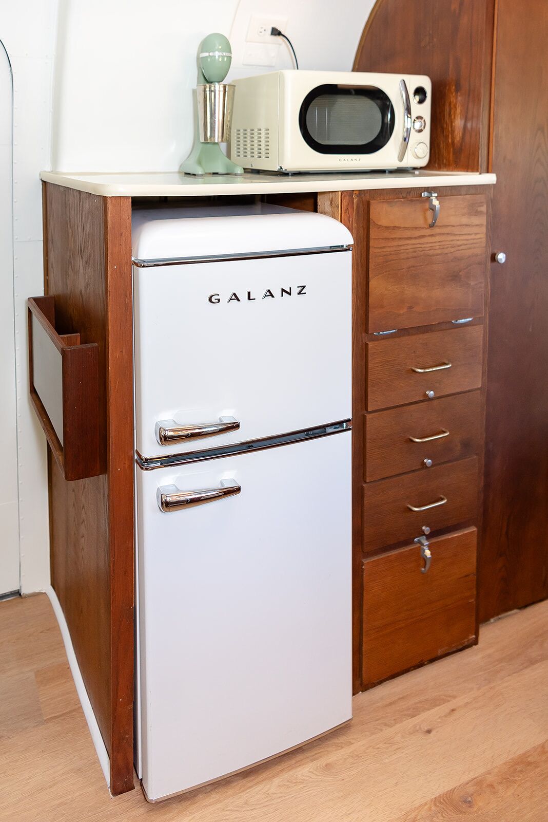 A white Galanz refrigerator with a microwave on top, a wooden cabinet with drawers, and a wooden door.
