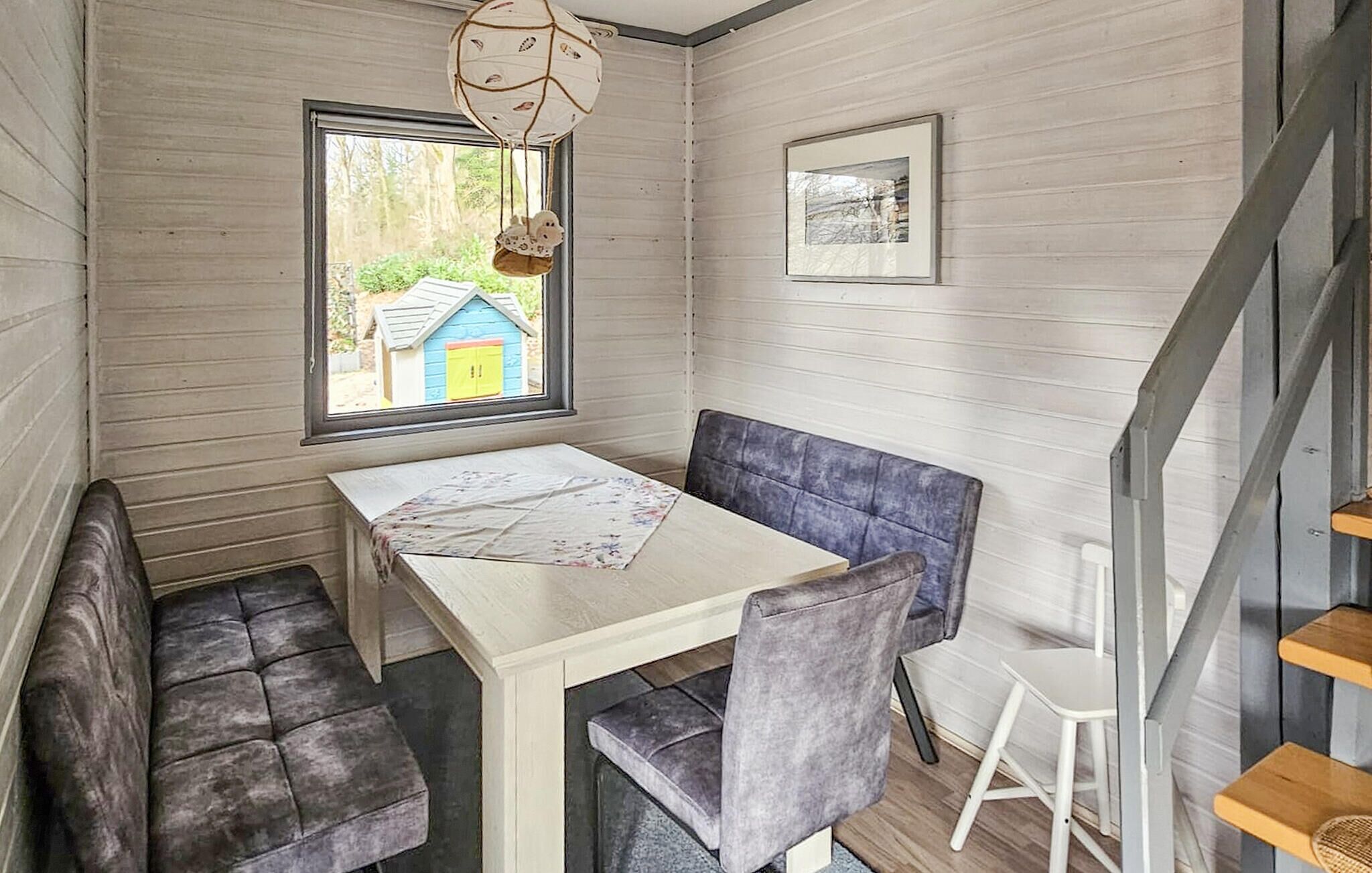 A room with a table and bench seating, a window with a view of a blue house, and a framed picture on the wall.