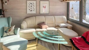 A living room with a sofa, armchair, and glass coffee table. There are framed pictures on the wall and a window with a view.