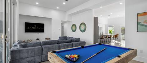 A modern living room with a pool table, grey sofa, and a dining area with chairs.