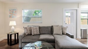 Ein Wohnzimmer mit einem grauen Ecksofa, einem Glastisch, einer Lampe und einem Fenster mit Jalousien.