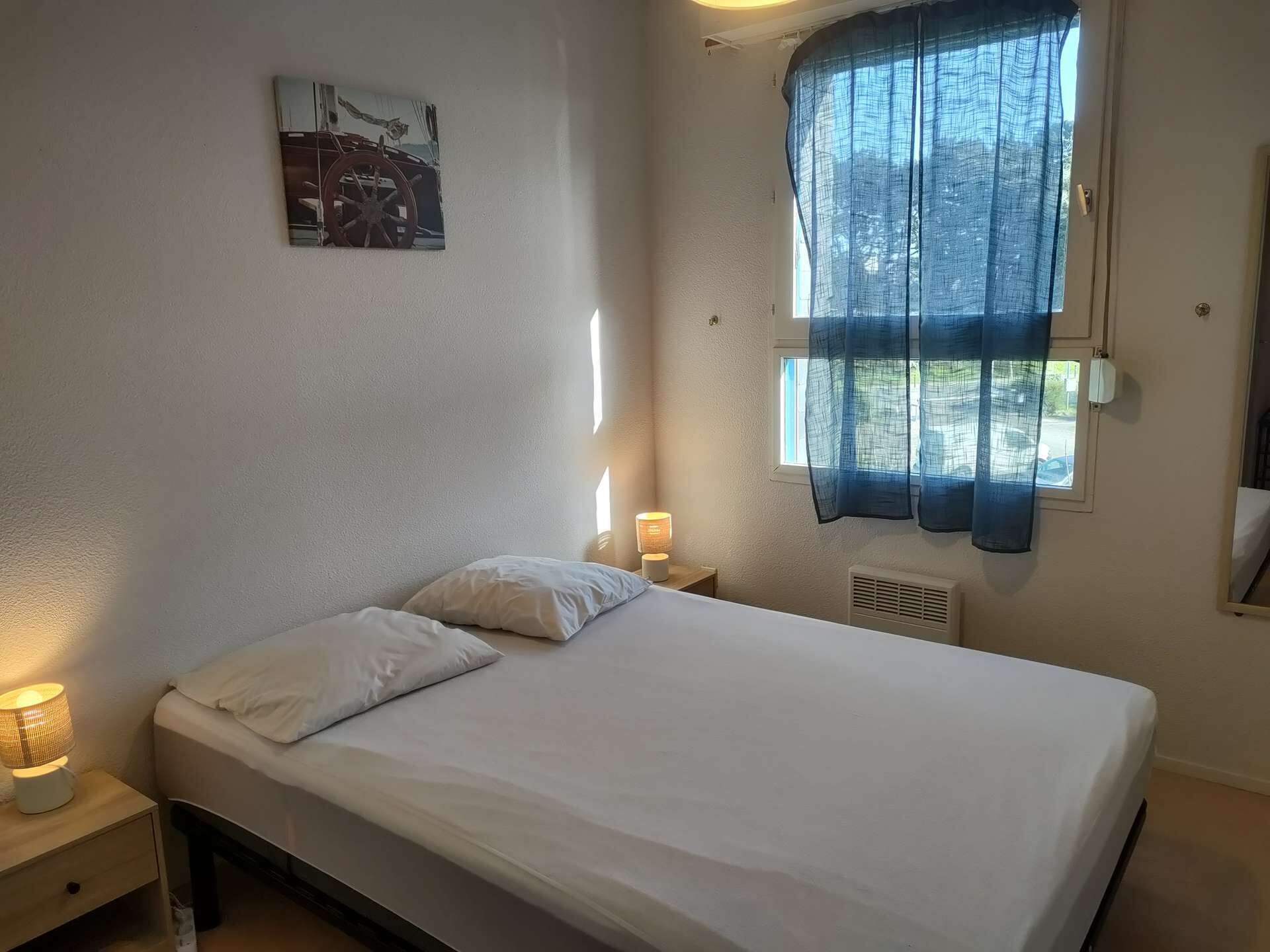 Un dormitorio con cama, mesita de noche con lámpara, ventana con cortinas e imagen colgada en la pared.