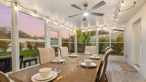 Un coin repas avec une table dressée pour quatre personnes, de grandes fenêtres, un ventilateur de plafond et une vue sur l’extérieur.