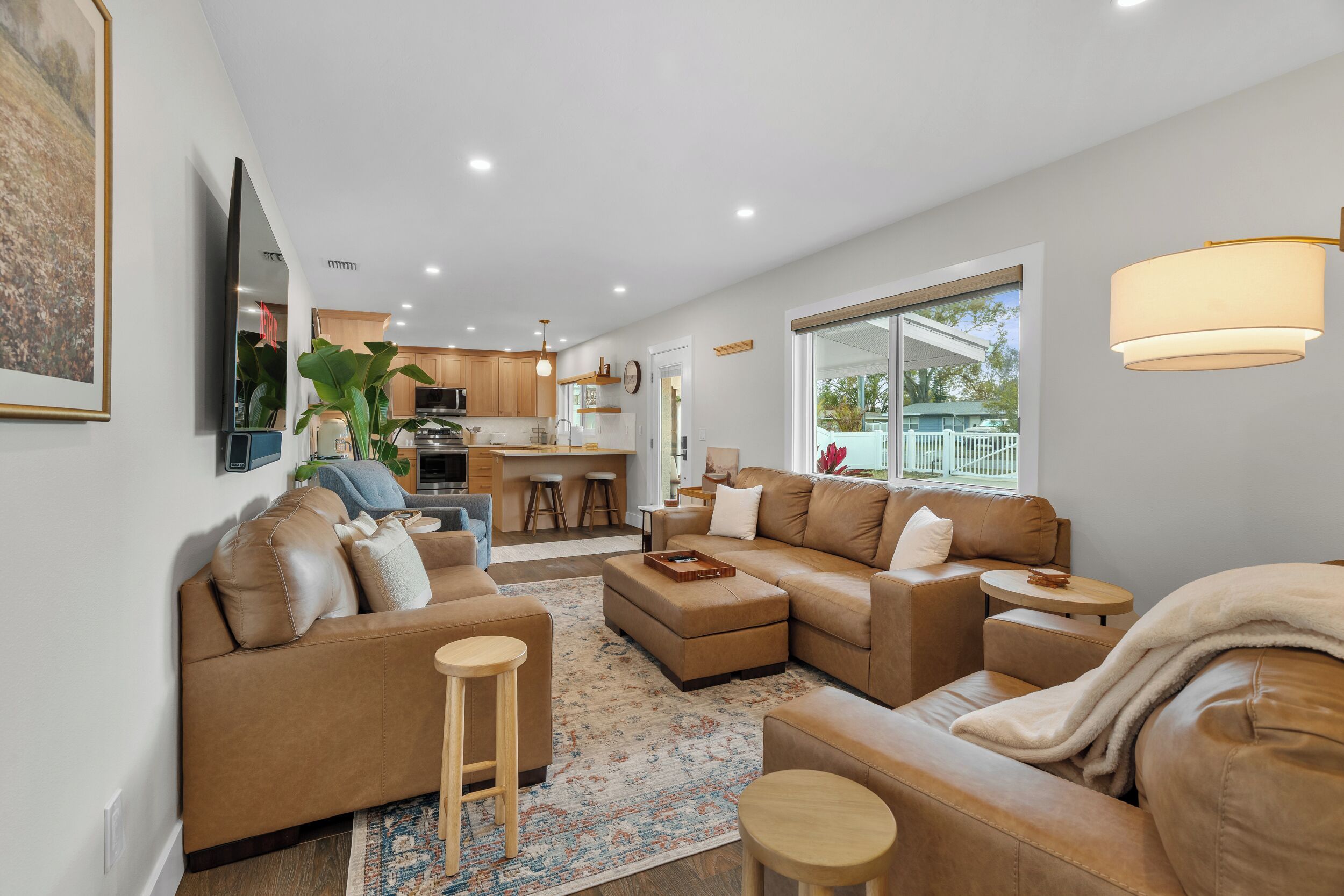 Ein modernes Wohnzimmer mit einer Couch, Hocker und einem Couchtisch. Im Hintergrund ist eine Küche mit Holzschränken und einem Essbereich zu sehen.