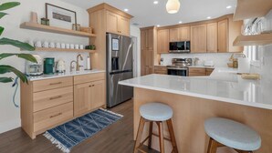 Une cuisine moderne avec des meubles en bois, un réfrigérateur en acier inoxydable, un îlot central doté de tabourets et un luminaire suspendu.