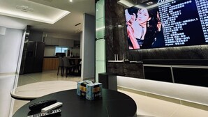 A modern living room with a large flat-screen TV mounted on a dark wall, a ceiling fan, recessed lighting, and a round coffee table with remote controls.
