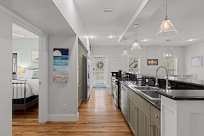 A modern kitchen with a central island, pendant lighting, and a view into a bedroom. - Magnolia Blue - 3BR/3BA Single-Story Cottage - Sleeps 8 - Near Community Pool (Inlet Beach)