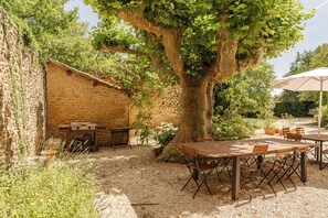 An outdoor patio with a stone wall, a wooden table, and a large tree providing shade. - Maison Margerie - Provencal farmhouse with swimming pool and view of Mont Ventoux (Colonzelle)
