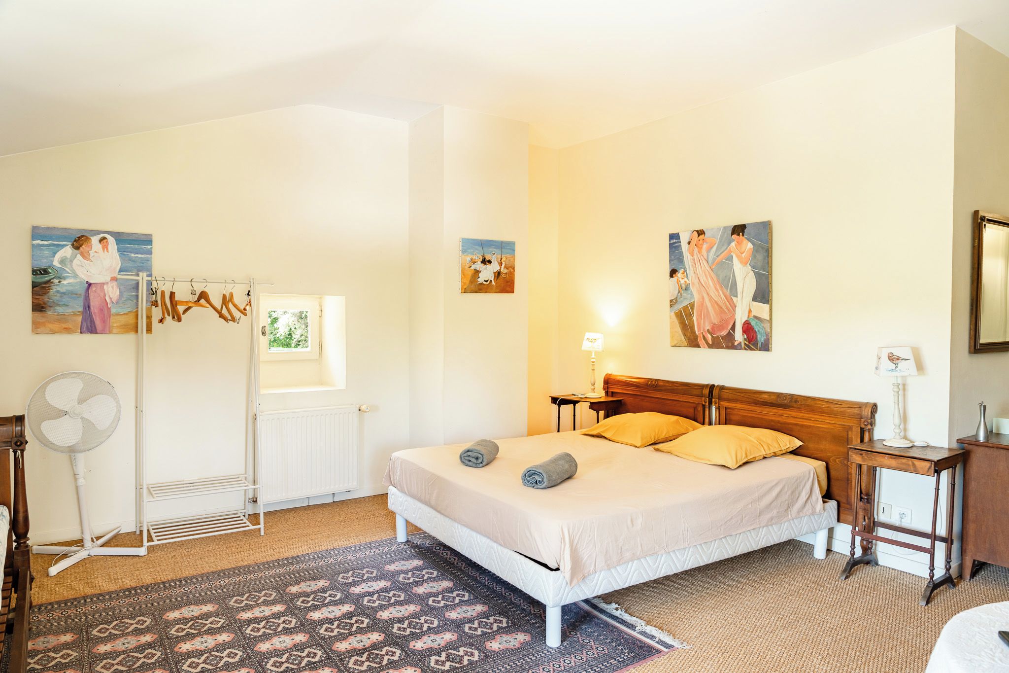 Une chambre à coucher avec un lit, un radiateur, une fenêtre, un ventilateur et des tableaux accrochés aux murs.