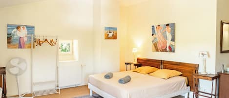 Un dormitorio con una cama, un radiador, una ventana, un ventilador y cuadros en las paredes.