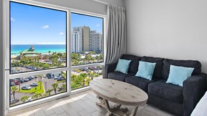 A room with a large window, a black sofa with blue cushions, and a wooden table. - Luau II 6633 (Miramar Beach)
