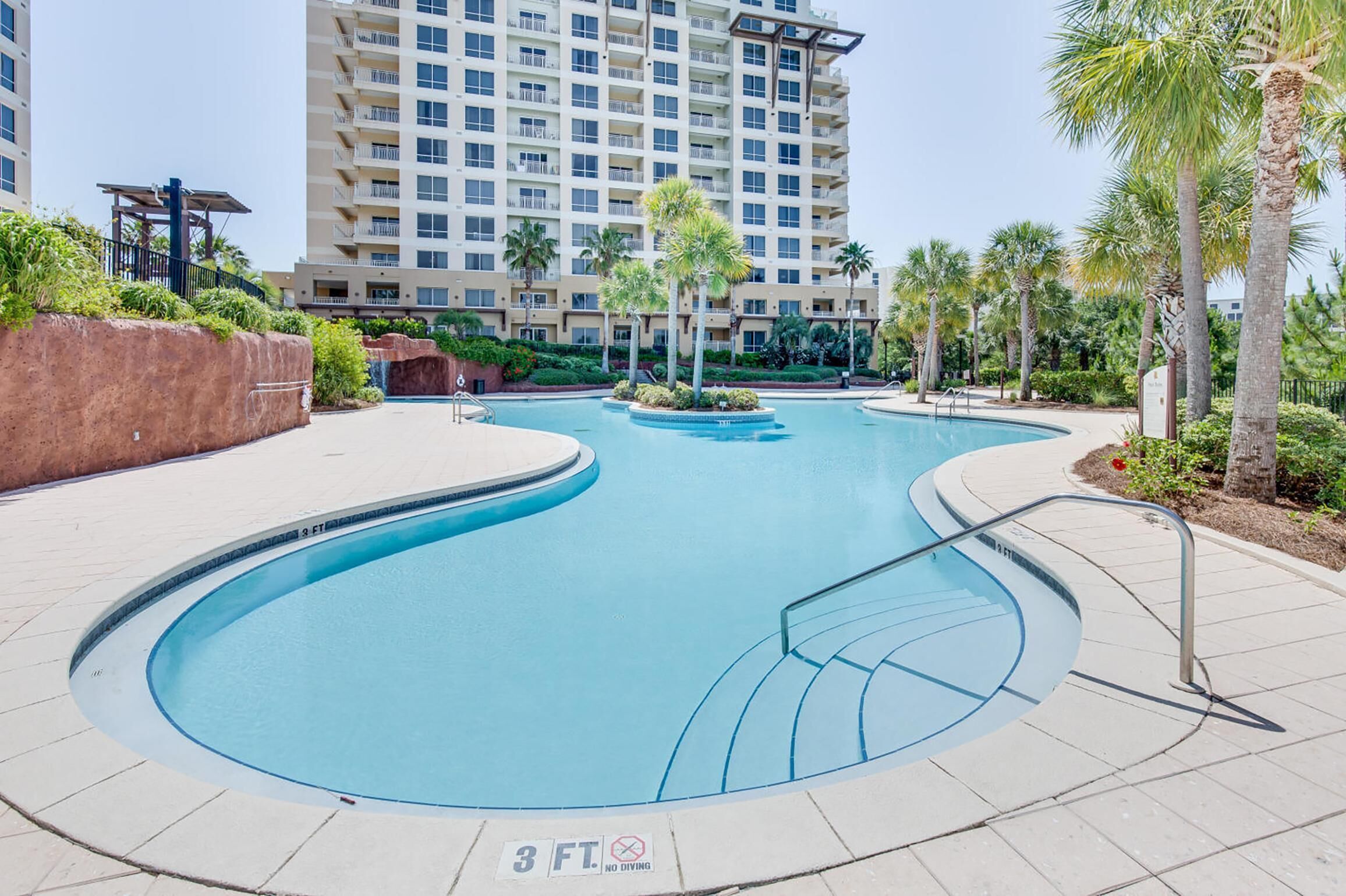 A swimming pool with a 3 ft depth limit sign, surrounded by palm trees and a multi-story building in the background.