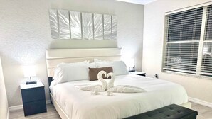 Una habitación de hotel con una cama grande, un reposapiés negro, una mesita de noche con lámpara y una ventana con persianas.
