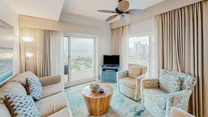 A hotel room with a sofa, armchair, TV, and a balcony with curtains. - Luau II 6832/6834 - Sunset Suite (Miramar Beach)