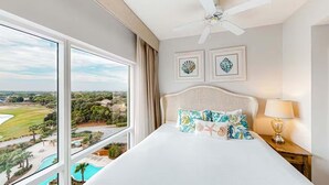 A hotel room with a large bed, a ceiling fan, framed artwork, a nightstand with a lamp, and a view of a pool and landscape. - Luau II 6832/6834 - Sunset Suite (Miramar Beach)