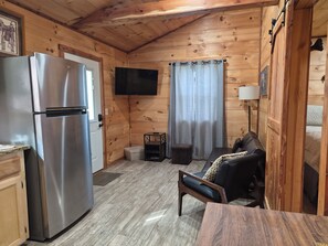A compact kitchen with a stainless steel refrigerator, a flat-screen TV mounted on the wall, a wooden dining table, and a sofa in a cabin-style room with wooden walls and floors. - Get away from it all in this beautiful Paradise in Northwest Arkansas! (Kingston)