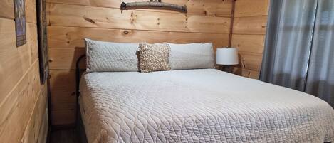 Una habitación de cabaña de madera con cama, cabecera, lámpara y cortina.
