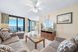 A spacious living area with a ceiling fan, a flat-screen TV, and a view of the ocean through large windows. - Phoenix VII 911 | Beachfront Views | Indoor, Outdoor Pools | My Beach Getaways (Orange Beach)