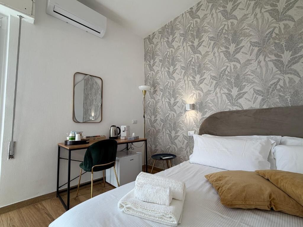 A hotel room with a bed, a desk, a chair, a small table, a mirror, and a wall with a leaf pattern.