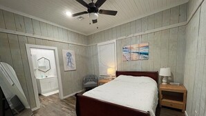 Een slaapkamer met houten vloer, een bed, nachtkastje, stoel, plafondventilator en kunstwerken aan de muur.