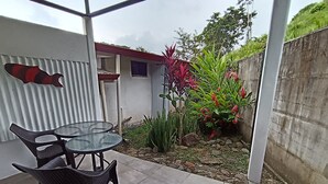 A patio with a glass table and chairs, surrounded by a garden with various plants and a small building in the background. - Cute studio with AC near Chaman beach Uvita Puntarenas (Uvita)