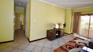 Una habitación con paredes amarillas, piso de baldosas, un televisor sobre un mueble de madera, una ventana con cortinas y una puerta que da a otra habitación.