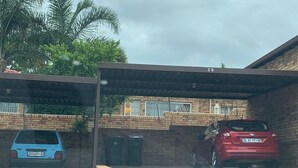 Un área de estacionamiento techada con dos autos, un edificio de ladrillo y una palmera al fondo.
