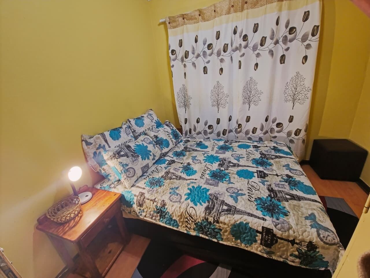 A bed with a floral and architectural print, a bedside lamp, and a small wooden nightstand with a lamp and a decorative item.