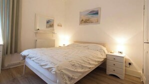 Quarto com cama, mesas de cabeceira, radiador e duas imagens emolduradas na parede.