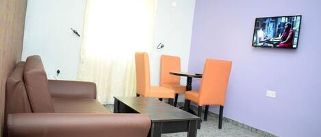 Une pièce avec un canapé marron, deux fauteuils orange, une petite table, un téléviseur fixé au mur et une fenêtre avec des rideaux.