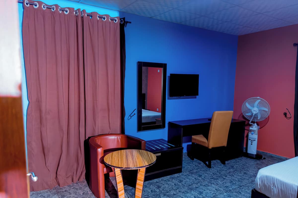 Hotelkamer met een bed, bureau, stoel, klein tafeltje, spiegel, televisie en een staande ventilator.
