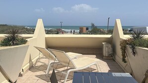 En balkon med to liggestole, et bord og udsigt til strand og hav.