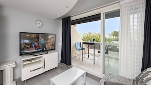 Un soggiorno moderno con televisione a schermo piatto, un tavolino da caffè bianco e un divano. È presente un balcone con tavolo e sedie, e vista sull'esterno.
