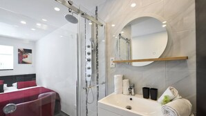Une salle de bain moderne avec une douche à parois de verre, un miroir rond, un lavabo blanc et un fauteuil de massage rouge.
