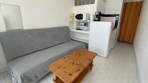 Um espaço de estar compacto com sofá cinzento, uma mesa de madeira, uma kitchenette com micro-ondas e máquina de café.