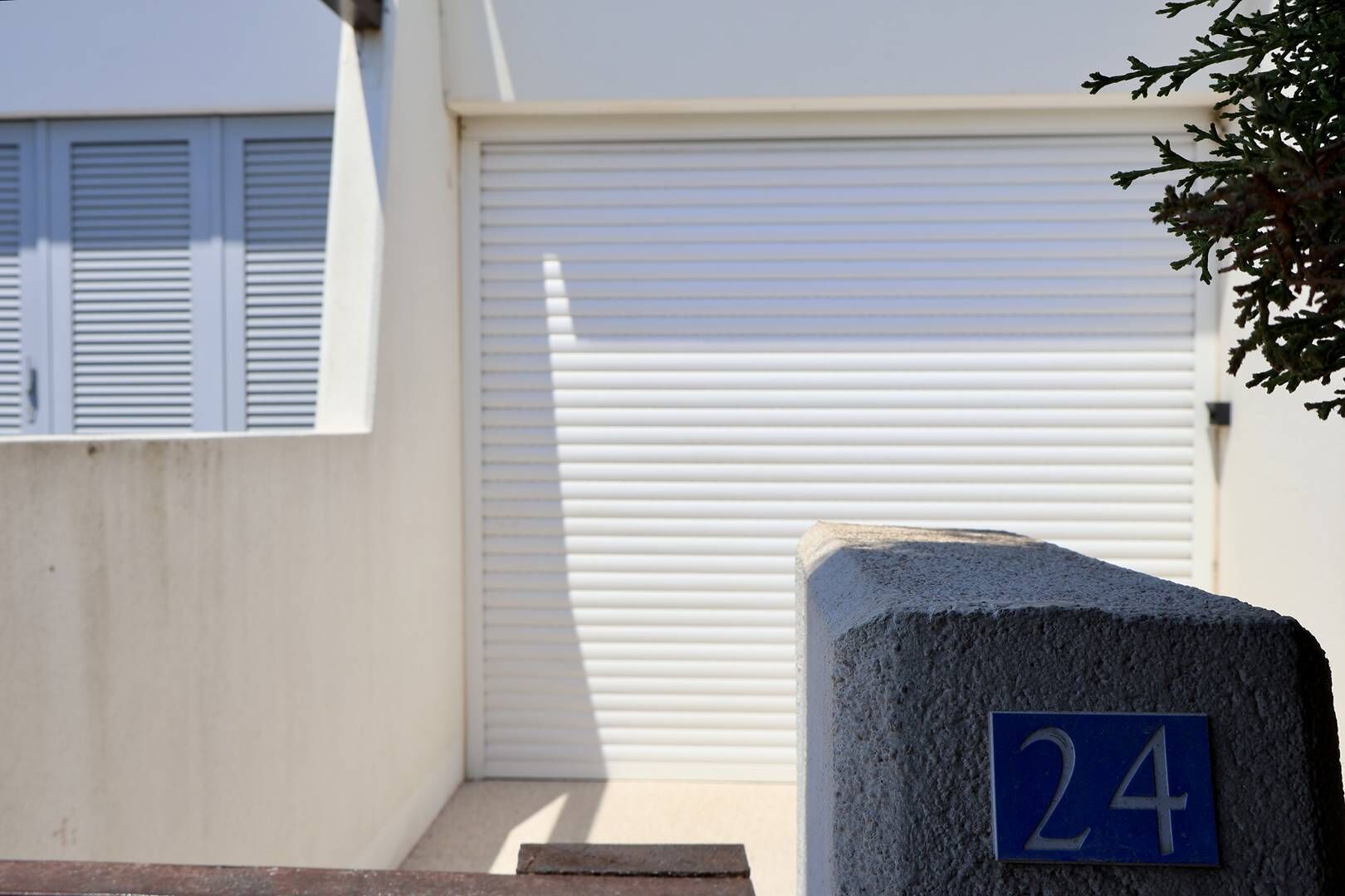 Un bâtiment avec une porte de garage fermée, un bloc de béton portant le numéro 24 et une fenêtre avec des stores horizontaux.