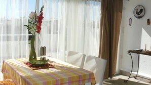 Un coin repas avec une table recouverte d’une nappe à carreaux, des chaises blanches, un vase avec des fleurs et une fenêtre ornée de rideaux légers.