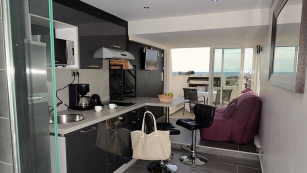 A modern kitchen with a built-in oven, a coffee machine, and a dining area with a purple chair and a view of the sea. - DIAMANT - Port Nature Apartment 81 - Luxury - 2 people (AGDE)