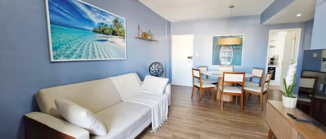 Una sala de estar con sofá, mesa de comedor, sillas, un ventilador y una cocina visible a través de una puerta abierta.