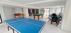 A room with a ping pong table, a pool table, and a billiards table. There are chairs and a small table with a green tablecloth. - COPBV0101 - Edificio Torres do Iguatemi (Salvador)
