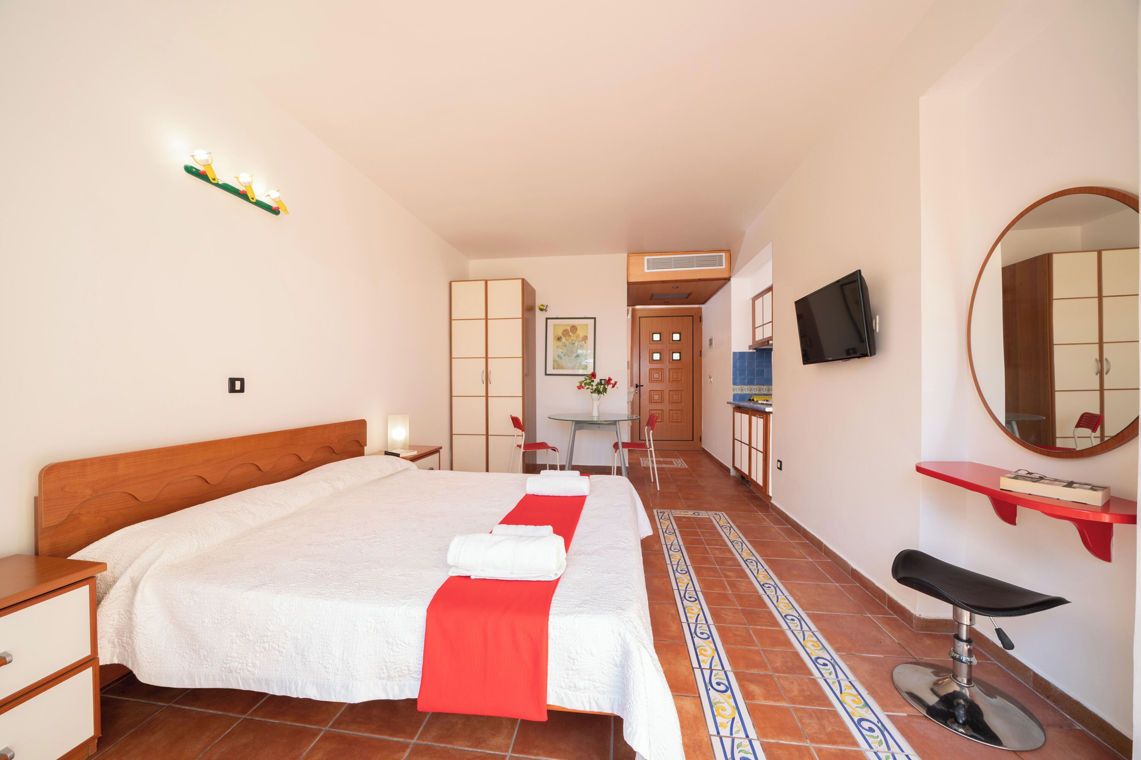 Habitación de hotel con una cama grande, zona de estar con mesa y sillas, televisión, espejo y baño con lavamanos y silla.
