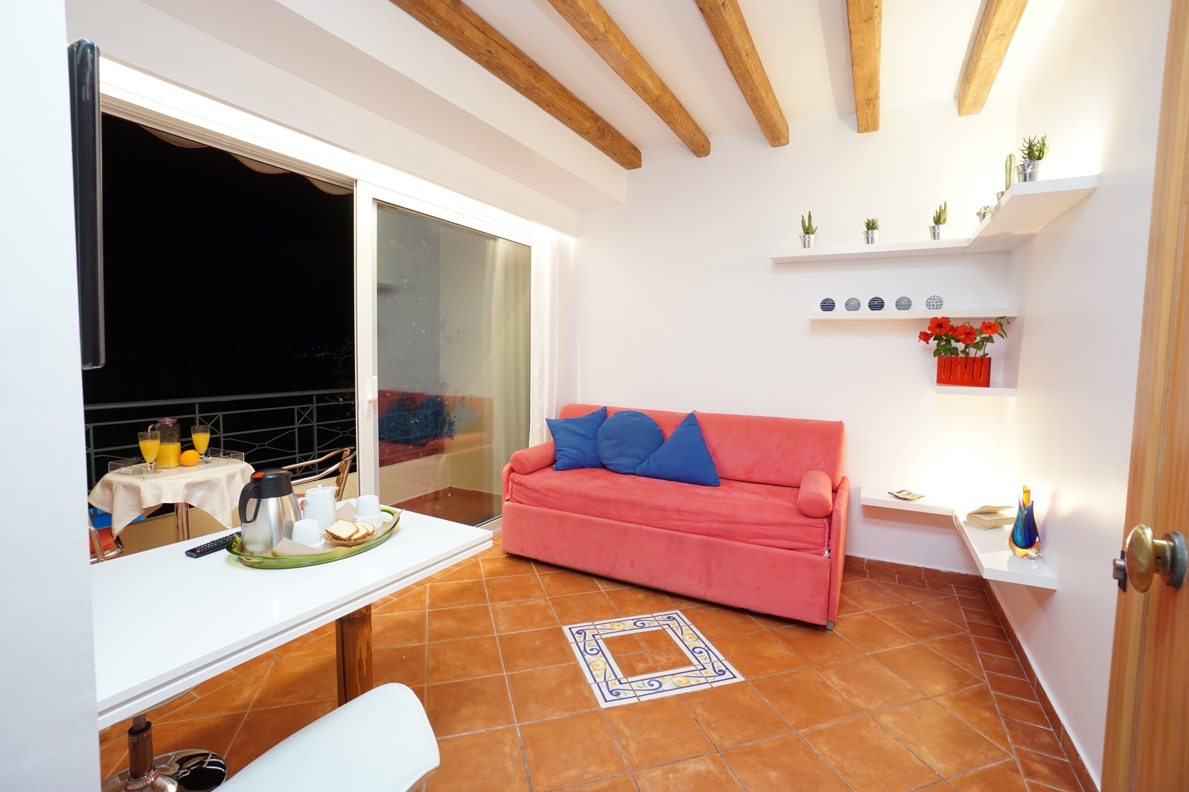 A room with a red sofa, a white table with breakfast items, a window, and a tiled floor.