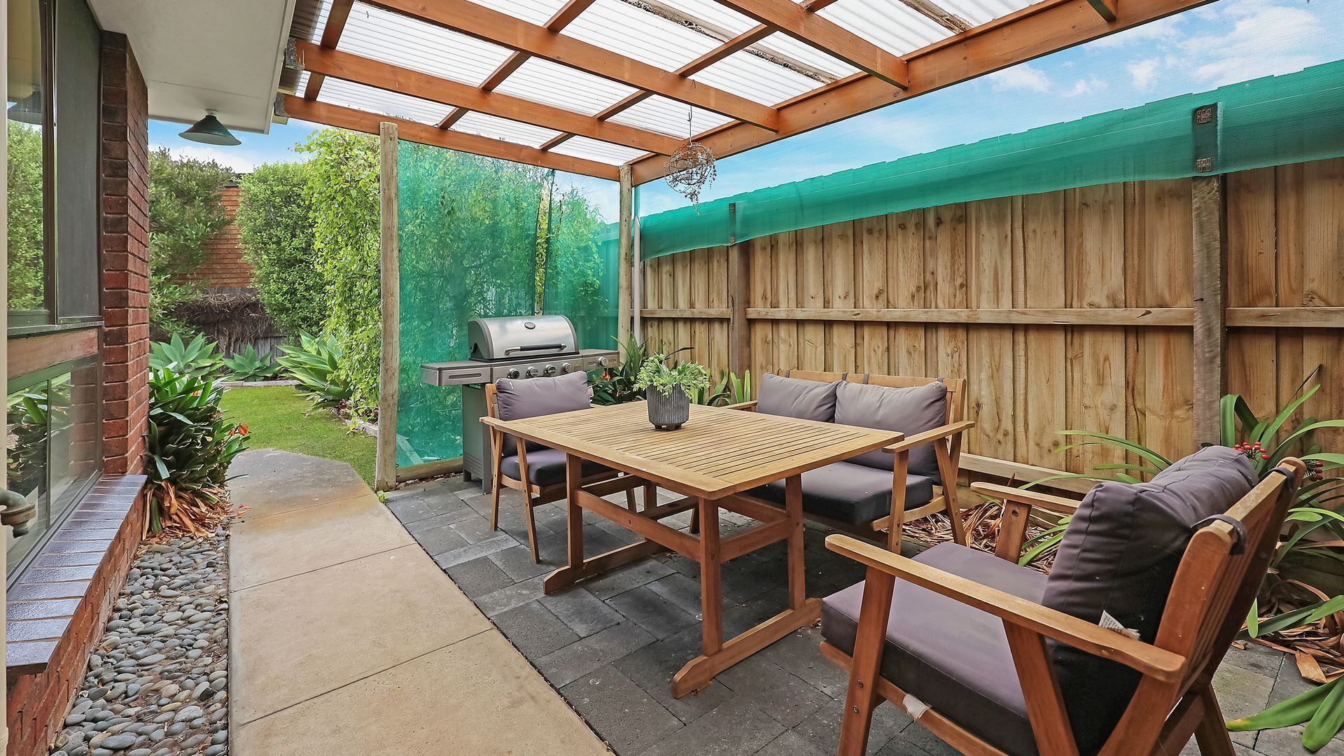 Un patio con mesa y sillas de madera, una parrilla y un área cubierta con vista al jardín.