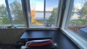 A room with a bay window overlooking a lake, a small table, and a bench. - Water Front Lake House - Black Lake, NY (Ogdensburg)