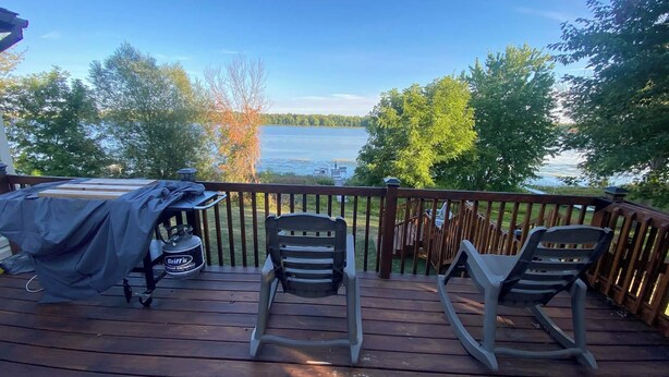 A wooden deck with two chairs, a grill, and a view of a lake. - Water Front Lake House - Black Lake, NY (Ogdensburg)