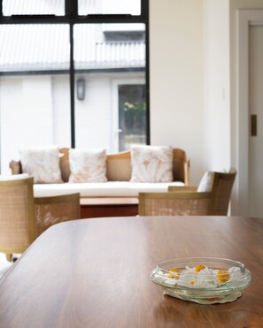 A wooden table with a glass bowl containing flowers. In the background, there is a sofa with patterned cushions and a window with blinds.