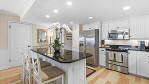 A kitchen with white cabinets, a stainless steel refrigerator, a microwave, and an island with chairs. - Sandpiper 514 (New Smyrna Beach)