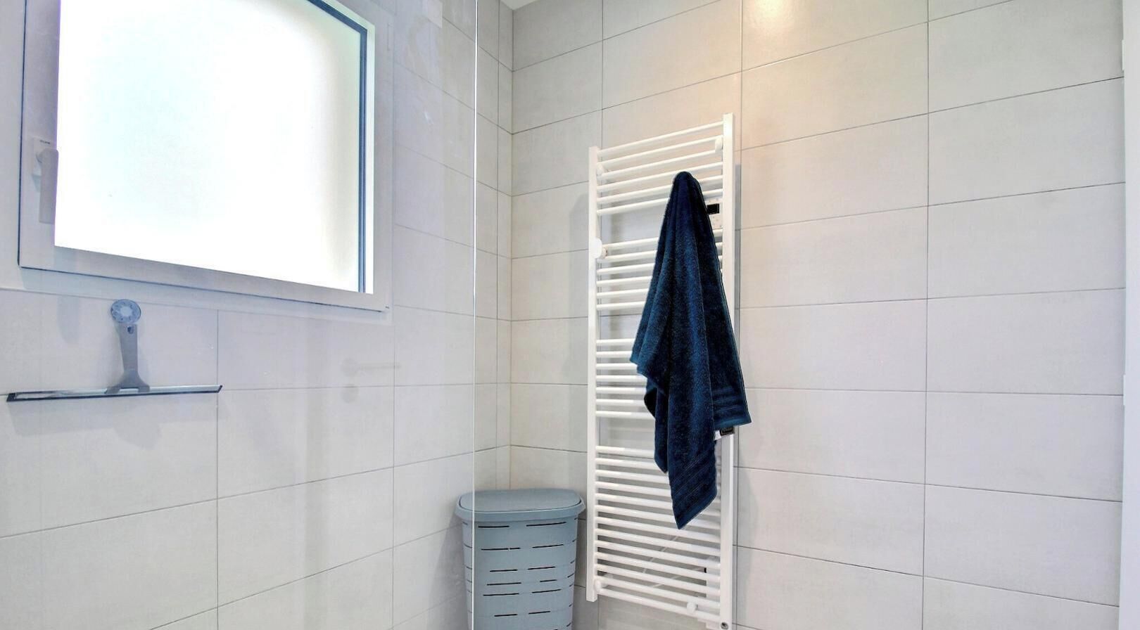 A bathroom with white tiled walls, a white towel rack, a blue trash bin, and a window with blinds.